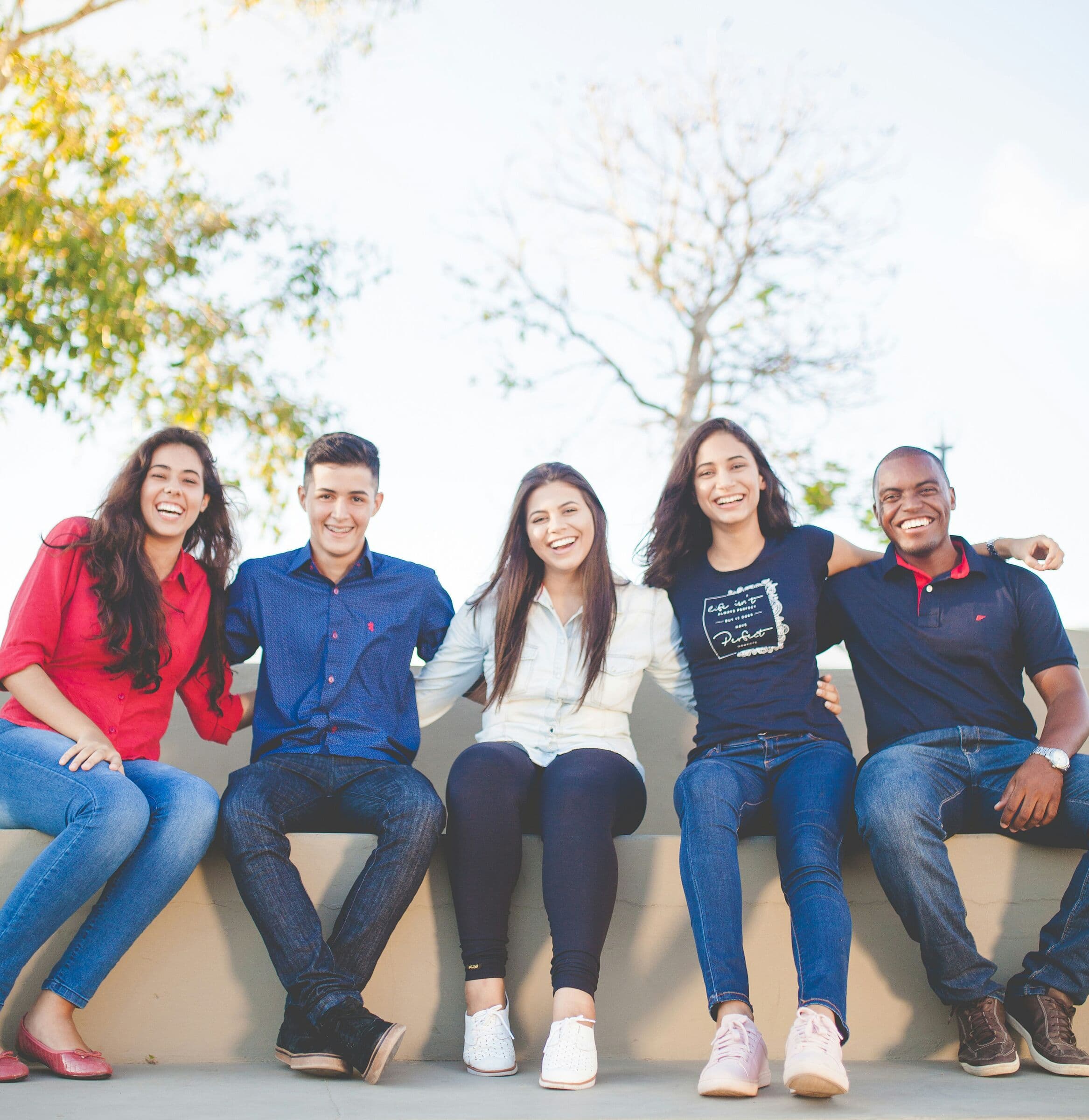 Five smiling young people sitting side by side on a low wall outdoors